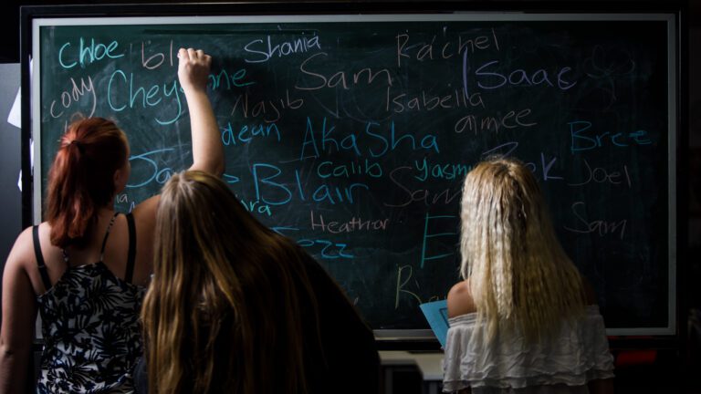 Three girls write on chalk board.